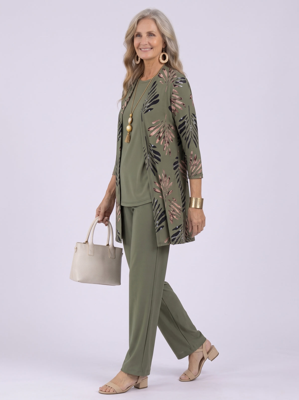 Donna con completo verde oliva in tre pezzi: cardigan lungo a foglie nere e rosa, top girocollo e pantalone ampio, in movimento